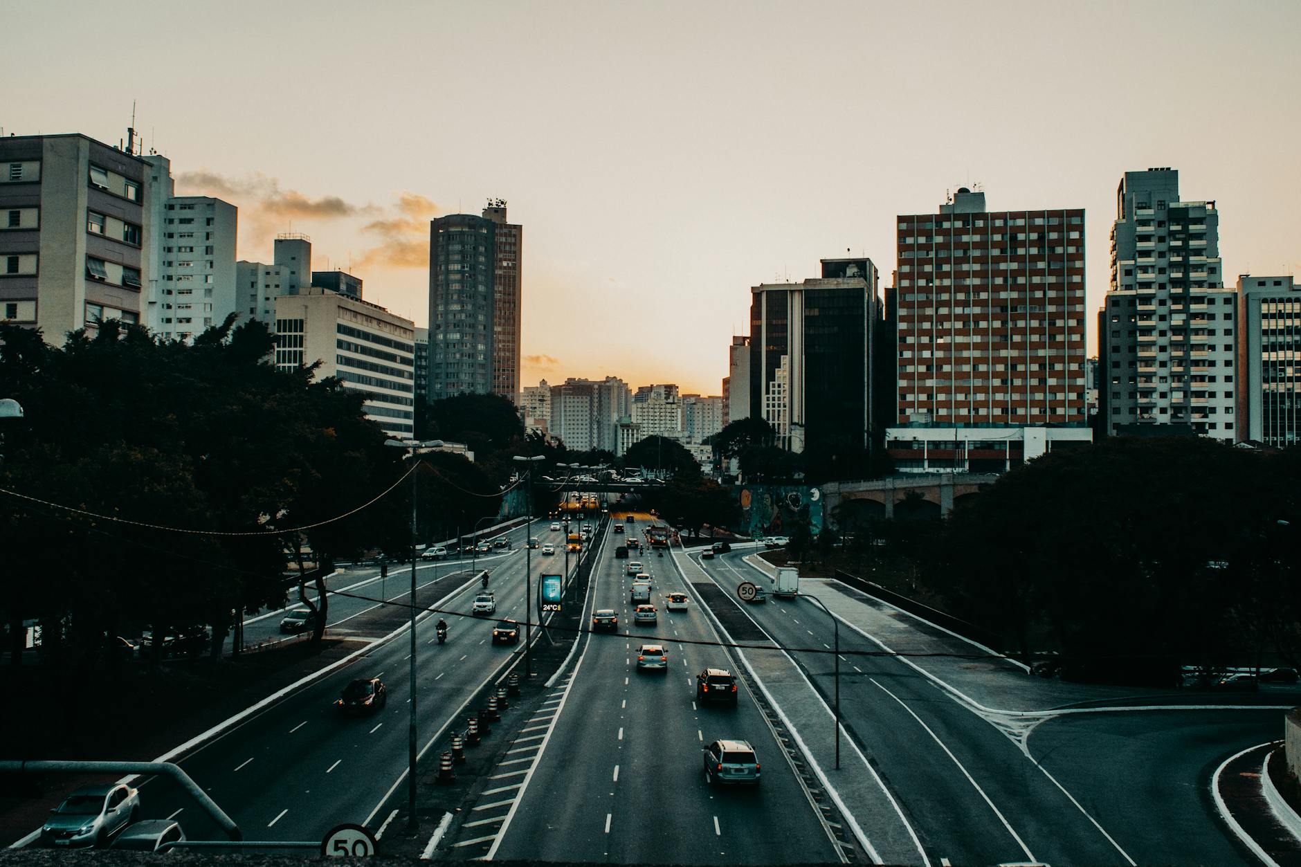 Various vehicles riding along asphalt road near modern buildings in city district against sunset sky